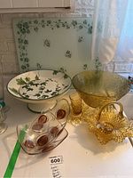 Overview photo showing several glass items including amber glass bowls and baskets, a white pedestal bowl and rectangular tray with green leaf decoration, and a clear decorative bowl with swirl patterns.