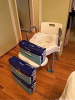 Photo showing white plastic commode chair with grey armrests, adjustable legs, and three unopened packs of adult diapers stacked on and beside the chair.