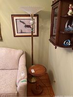 Full view of torchiere lamp with frosted glass shade and metal base next to beige upholstered sofa, with wooden round pedestal table beside it.