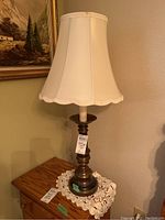 Photo of one brass lamp on wood side table with white fabric lampshade, showing turned base and scalloped edge shade.