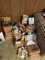 Wide view of multiple boxes of various household lightbulbs, electrical outlets, and extension cord on a floor next to wall and couch