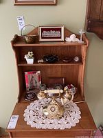 Wide shot showing vintage flower-embellished rotary phone on lace doily, with framed religious images, candles, small decorative bowls and basket on wooden shelf behind.