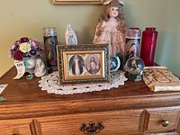 A group photo showing religious candles, figurines, and a porcelain doll on a wooden dresser with a white doily underneath.