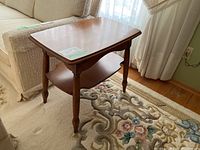 First wood end table showing rectangular top, turned legs, and lower shelf, placed on a floral patterned rug next to a cream upholstered couch.