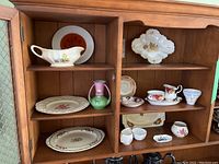 Photo showing wooden cabinet shelves filled with assorted porcelain tableware including teacups with saucers, platters, creamer, small pitcher, and plates.
