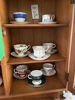 Full view of eight assorted teacups and saucers displayed on wooden shelves inside a cabinet