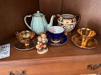 Display of all seven items including teapot, three teacups with saucers, Sadler pitcher, and Hummel figurine