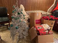 4 ft silver tinsel Christmas tree decorated with green spherical ornaments next to a 6 ft white vinyl Christmas tree and several boxes containing holiday decorations and artificial wreaths.