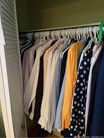 Closet rod filled with multiple women's blouses and tops hanging on white hangers, colors including white, light blue, yellow, black, and blue with polka dots.