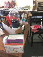 Photo showing various items including a cardboard box containing glass globe displays, greeting card box, and packaged molds, arranged on chairs and tables.
