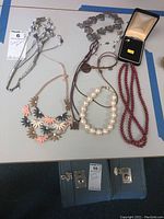 Photo showing seven items of costume jewelry arranged on a table, including multiple necklaces, a bracelet, and single pearl earring in box.