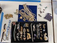 Full view of the jewelry tray with assorted bracelets, earrings, brooches, pearl necklaces, and two separate groups of earrings and pins on the white table.