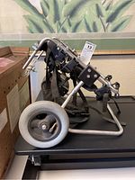 Side view of small dog mobility cart showing aluminum frame, large rear wheels, harness, and adjustment components.