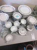 Full view of multiple stacks of white porcelain plates of various sizes, cups and saucers, cream and sugar bowl with lid, and serving pieces all decorated with a silver floral and vine trim pattern.