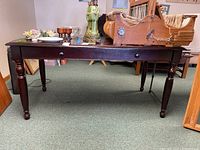 Front view of the large wooden office desk showing the full desk, turned legs, and single drawer with a round knob. Visible scratches and wear marks on the surface.