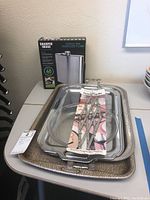 Original lot photo showing Sharper Image engraved flask in unopened box, stacked metal serving trays, and packaged chrome table mat.