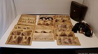 Photo of multiple stereoview cards arranged on a table, showing varying sepia tones and scenic images, alongside the binoculars and carrying case on the right.