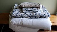 Stack of bedding items including white down filled duvet, folded reversible duvet covers with geometric pattern, and beige/tan bed skirt on wooden surface