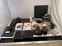 Photo shows an array of electronics and accessories including laptop chargers, iPad and iPhone cases, auxiliary cables, pedometers, alarm system parts, and DVDs.