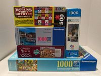 Stack of five puzzle boxes including two Ravensburger puzzles, one Thomas Kinkade, one Charles Wysocki, and one double-sided puzzle by Ceaco