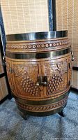 Front view of barrel-shaped liquor cabinet with carved grape clusters and leaves, brass handles, metal trim, and casters