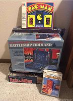 Photo showing stacked board game boxes including Pac-Man card game, Battleship Command electronic game, Axis & Allies board game, and Travel Battleship box.