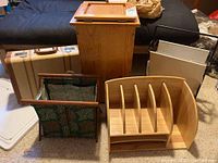Photo showing wooden magazine rack, fabric tote with wooden frame, vintage suitcase, wooden storage box, and part of a dark sofa or futon in background.