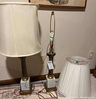 Pair of table lamps with white marble square bases, vertical brass fluted columns, and one lamp fitted with bulb and harp. Two white fabric lamp shades placed beside lamps.