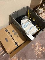 Plastic bin with black curved road racing track pieces, yellow plastic supports, folded instruction sheets, and small bags inside. Cardboard boxes on floor next to bin.