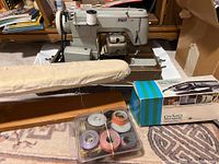 Overall view of Pfaff 18 sewing machine with thread spool, Dymo Deluxe Tape Writer box, small ironing board, and sewing threads