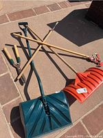 Photo showing gardening tools laid on patio including a red snow shovel, flat shovel with green handle, pole saw with red blade, and green-handled loppers