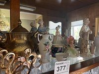 Shelf with vintage ceramic figurines including women and children in dresses, metalware such as teapots, pitchers, swan napkin holders, and ceramic cups and saucers.