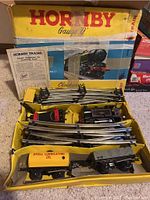 Photo showing Hornby box with multiple metal train tracks, black locomotive, gray coal tender, yellow Shell oil boxcar, and grey boxcar inside, placed on carpeted floor.