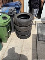Stack of four used all-season tires placed outdoors next to trash bins.