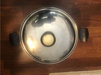 Top view of a large stainless steel cooking pot with a clear lid that has a black handle knob in the center and black handles on the pot sides.