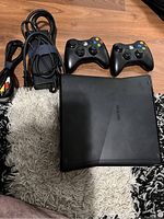Xbox 360 console with two wireless controllers and power/AV cables laid on a rug.