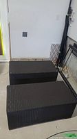 Two black woven rattan rectangular patio tables or benches placed side by side against a white wall in a garage or storage area.