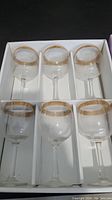 Set of six clear drinking glasses with gold decorative trim around the rims, arranged in original white box packaging.