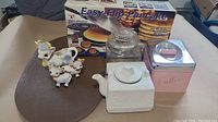 Photo showing pancake maker box, three ceramic cow figurines, white ceramic tea kettle, clear glass canister, pink metal coffee canister, and one brown placemat under them.