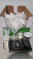 Full view of lot showing several clear crystal glasses in cardboard box, black rectangular plates with gold floral design, small white dietary scale, and small dishes including a white bowl with blue rim.