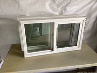 Two white vinyl framed basement slider windows with sliding glass panes and attached screens shown stacked on a table, viewed from above and side, showing use and dirt.