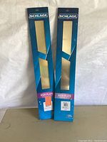 Lot of two bright brass door kick plates in original blue Schlage packaging standing upright on a beige surface, showing front of packages with product and brand name.