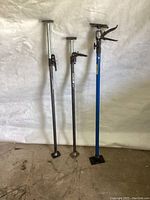 Three adjustable metal cargo bars of varying heights with rubber ends and locking mechanisms, one painted blue with visible labels.