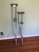 Full view of two different pairs of aluminum crutches standing against a wall, showing their general condition and construction.