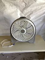 Front view of gray Lasko box fan with white blades and power cord visible, placed on a table.