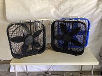 Four square box fans on a white table, black, white, and blue color, missing feet, visible dust and dirt, two control knobs on top of each fan.
