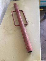 Red metal T post driver hammer lying on a beige table, showing full length and T-shaped handles.