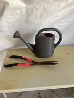 Photo showing the watering can and clippers placed on a beige surface, capturing the full items.
