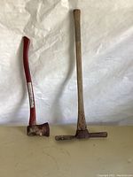 Photo showing the full lengths of the ax and pick, placed side by side on a surface with a white backdrop.