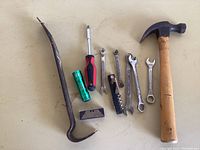 Claw hammer with wooden handle, pry bar, screwdriver, small flashlight, razor blade, multi-tool with corkscrew, and multiple wrenches arranged on a beige surface.